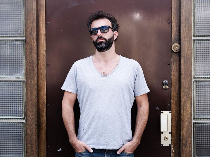 Simon Zaoui, debout devant une porte en bois, lunettes de soleil, t-shirt gris