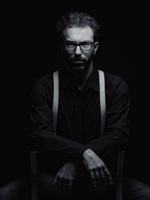 Simon Zaoui, en noir et blanc, assis sur une chaise, bretelles, regard face caméra