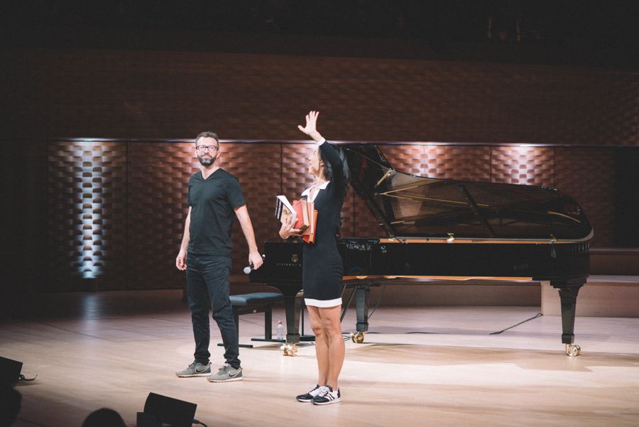 Sabine Quindou et Simon Zaoui saluant sur la scène de l’Auditorium, Sabine un bras levé et des livres sous le bras, Simon un micro à la main devant le piano à queue